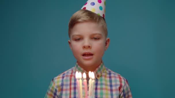 Pequeño cumpleañero con sombrero de fiesta soplando velas de cumpleaños y pidiendo un deseo. Niño feliz en el sombrero de cumpleaños sopla velas luces sobre fondo azul. — Vídeo de stock