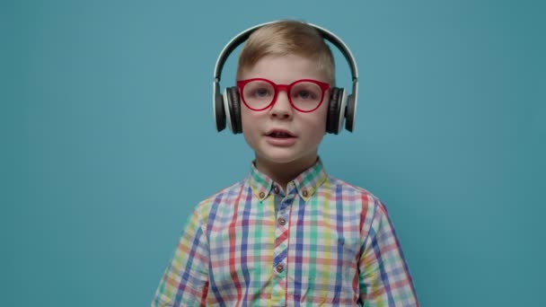 Menino pré-escolar em óculos e fones de ouvido conversando com câmera em fundo azul. Webcam vista do aluno falando olhando para a câmera. — Vídeo de Stock