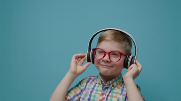 Cute young boy enjoys listening the music in headphones dancing on blue background in slow motion. — Stock Video