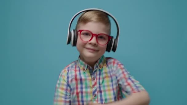 Симпатичный школьник в очках и наушниках танцует на синем фоне в замедленной съемке. Счастливый ребенок смотрит в камеру. — стоковое видео