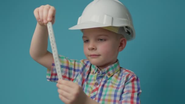 Menino pré-escolar em construção capacete branco segurando fita métrica em mãos sobre fundo azul. Criança sonhando em ser engenheiro arquiteto construtor. — Vídeo de Stock