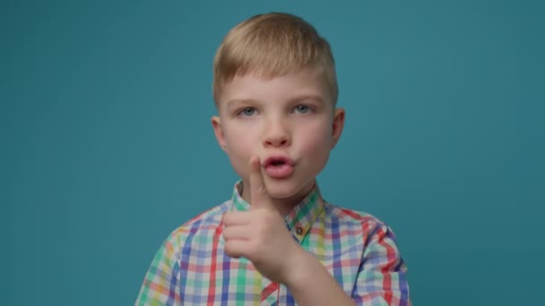 Muchacho preescolar mostrando el signo de silencio con el dedo índice por la boca de pie sobre fondo azul. Niño lindo pedir a estar en silencio con gesto de silencio. — Vídeos de Stock