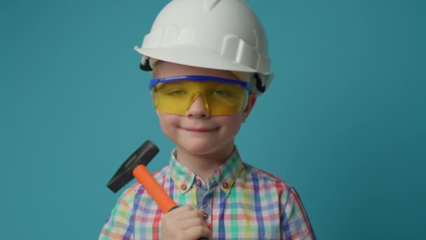 Mladý chlapec nosí bílý hardhat a ochranné brýle drží hummer v rukou s úsměvem při pohledu na kameru na modrém pozadí. — Stock video
