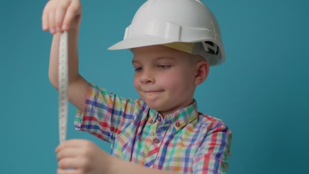 Kid constructor en protector blanco hardhat celebración cinta métrica en las manos sobre fondo azul. Niño soñando con ser ingeniero arquitecto. — Vídeos de Stock