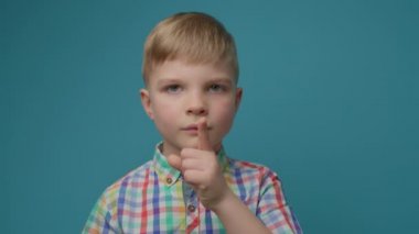 Mavi arka planda işaret parmağıyla sessiz hareketler yapan sevimli bir çocuk. Sarışın çocuk portresi sessiz ve sessiz bir işaret parmağıyla.