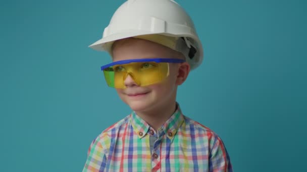 Garoto sorridente vestindo chapéu branco e óculos protetores amarelos segurando hummer em mãos no fundo azul. Jovem garoto engenheiro construtor. — Vídeo de Stock