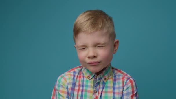 Niño rubio preescolar con los ojos cerrados de pie sobre fondo azul mirando a la cámara. 5 años de edad retrato de niño. — Vídeo de stock