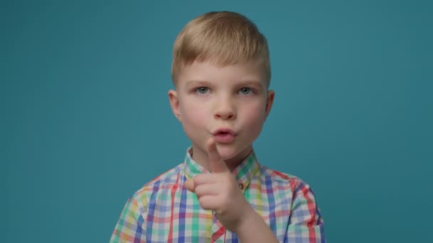 Enfant mignon faisant geste de silence avec index par la bouche sur fond bleu. Portrait de garçon blond montrant le silence et signe calme avec le doigt. — Video