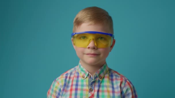 Koruyucu gözlük takan, sarı kask takıp kameraya bakan sevimli anaokulu çocuğu. Mavi arka planda genç bir inşaatçı. — Stok video