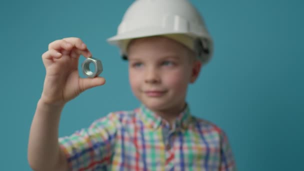 Kid worker usando capacete branco segurando parafuso porca nas mãos sobre fundo azul. Menino pré-escolar como engenheiro e construtor. — Vídeo de Stock