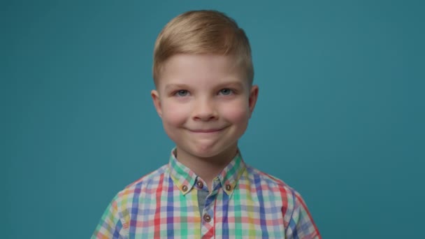 Lachende kleuterschool blonde jongen in kleur t-shirt kijken naar camera op blauwe achtergrond. 5 jaar oud kinderportret. — Stockvideo