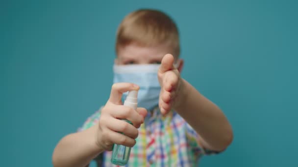 Kid bär ansikte skyddsmask med hjälp av handdesinfektionsmedel stående på blå bakgrund. Var socialt ansvarsfull. Stoppa spridningen av sjukdomar. — Stockvideo