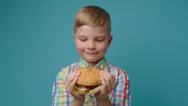 Egy óvodás fiú hamburgert tart a kezében, és a kék háttérben álló kamerát nézi. Boldog éhes gyerek élvezze a hamburgert kézzel.. — Stock videók