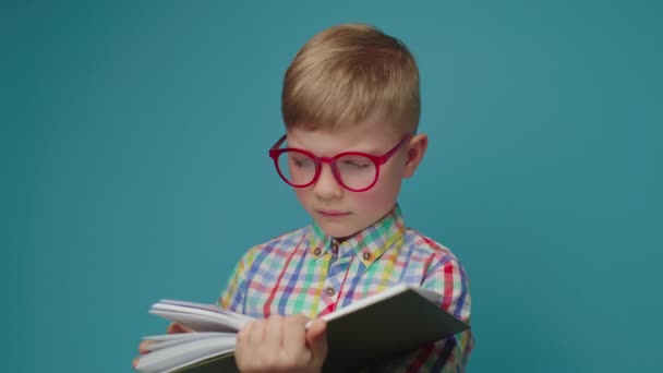 Upprörd skolpojke i glasögon läsa bok stående på blå bakgrund. Ungen gillar inte att läsa böcker.. — Stockvideo