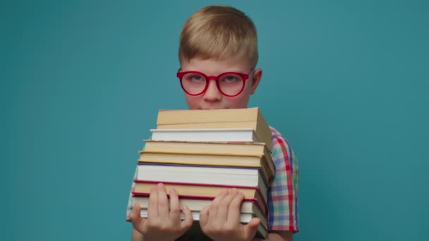 Μαθητής με γυαλιά που κρατάει βιβλία και χαμογελάει κοιτώντας την κάμερα πάνω σε μπλε φόντο. Έξυπνο παιδί που απολαμβάνει να διαβάζει βιβλία.. — Αρχείο Βίντεο
