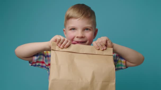 Miúdo faminto segurando caixa de entrega de papel com comida olhando para a câmera em pé no fundo azul. Menino animado sobre saco de entrega de fast food. — Vídeo de Stock