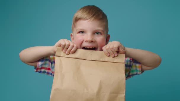 Счастливый ребенок держит бумажный пакет с едой, смотрящей в камеру на синем фоне. Мальчик взволнован коробкой быстрого питания. — стоковое видео
