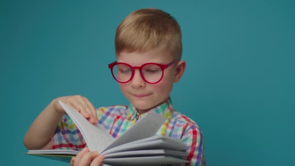 Дошкільний хлопчик в окулярах читає паперову книгу, тримаючи підручник в руках, що стоїть на синьому фоні. Щасливий школяр навчається з книгою . — стокове відео