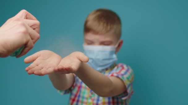 Mães mão usando higienizador de mão para criança vestindo máscara protetora de rosto em pé sobre fundo azul. Sê socialmente responsável. Parar a propagação de doenças. — Vídeo de Stock