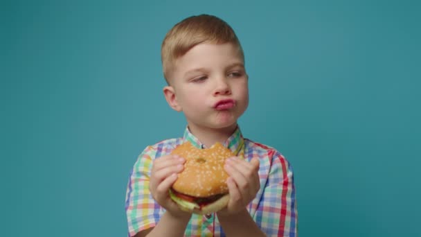 Blond kind dat hamburger eet in handen staand op een blauwe achtergrond. Pensive hongerige jongen geniet van fast food eten. — Stockvideo