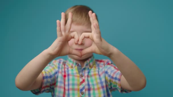 Ребенок держит руки в форме сердца, смотрит на камеру, стоящую на синем фоне. Символ "Сердце пальца", сделанный дошкольником. — стоковое видео