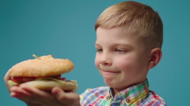 Detailní záběr hladové dítě jíst burger drží v rukou na modrém pozadí. Školka chlapec má rád fast food jíst. — Stock video