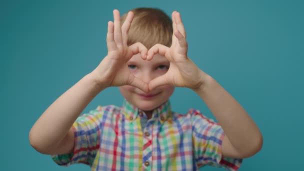 Niño sonriente haciendo forma de corazón con los dedos mirando a la cámara. Niño encantador sosteniendo el símbolo del corazón de la mano en frente de su cara. — Vídeos de Stock