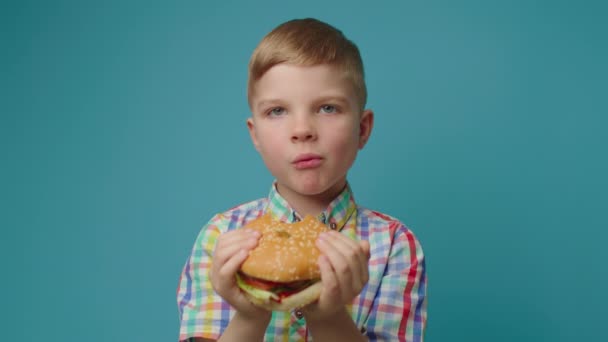 Roztomilý školák drží burger v ruce a jí lahodný cheeseburger stojící na modrém pozadí a dívá se na kameru. Kid žvýkání fast food. — Stock video