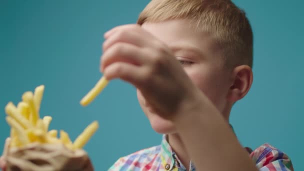 Close up of boy eating french fries with hands standing on blue background. Kid enjoys eating fried potatoes. — Stock Video