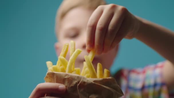 Närbild av barn hand tar bit pommes frites från papperspåse full av stekt potatis. Pojke äter pommes frites på blå bakgrund. — Stockvideo