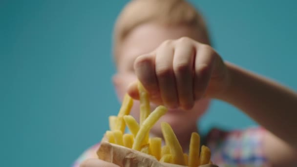 Крупным планом дети берут кусочек картошки фри из бумажного пакета, полного жареного картофеля. Мальчик ест картошку фри на голубом фоне. — стоковое видео