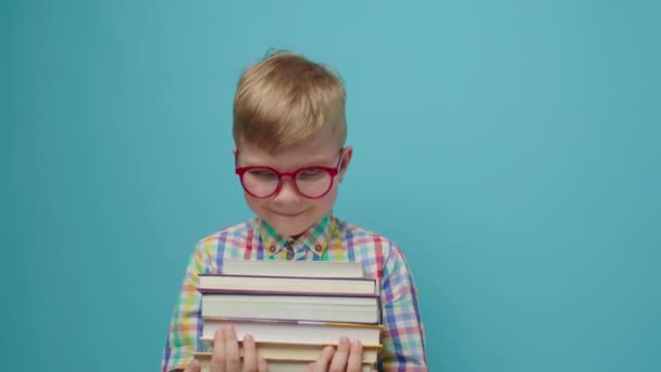 Preschool boy in eye glasses holding books and smiling looking at camera standing on blue background. Smart child enjoying reading paper books. — Stock Video