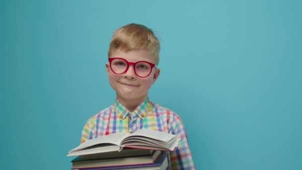 Красивый школьник в очках читает книгу и улыбается, глядя на камеру, стоящую на синем фоне. Умный ребенок любит учиться, читая бумажные книги. — стоковое видео