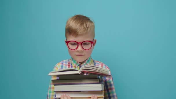 Smart skolpojke i glasögon läser pappersböcker och ler stående på blå bakgrund. Glad unge njuter av att läsa böcker. — Stockvideo