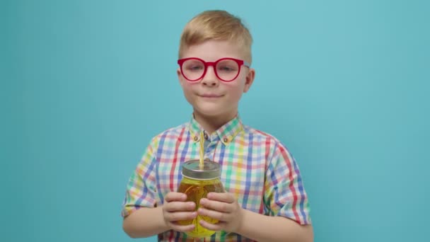 Дошкольник в очках пьет воду с соломинкой, держа бутылку на руках, улыбаясь, глядя на камеру, стоящую на синем фоне. Ребенок любит пить воду. — стоковое видео