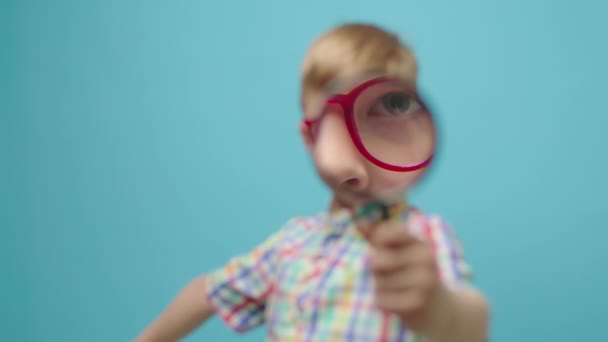 Der Vorschuljunge blickt durch eine Lupe in die Kamera, die auf blauem Hintergrund steht. Kind mit Brille durch die Lupe. — Stockvideo