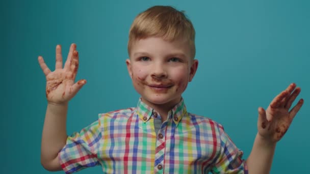 笑着男孩的脏巧克力脸给高五看着相机站在蓝色的背景。快乐的孩子喜欢巧克力. — 图库视频影像