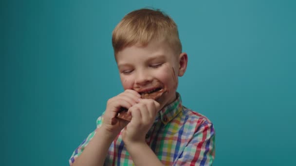 Niño feliz con la cara sucia comiendo barra de chocolate sosteniendo en las manos sonriendo a la cámara de pie sobre fondo azul. Chico alegre disfrutando de dulces de chocolate. — Vídeos de Stock