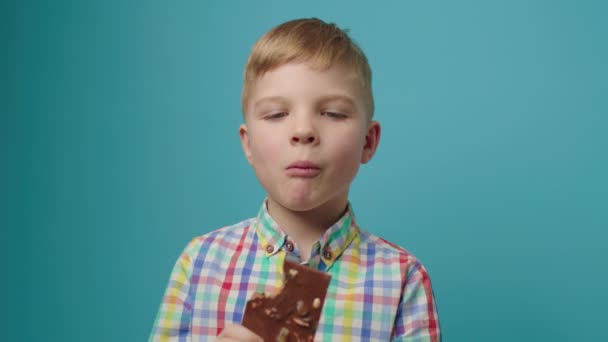 Šťastný chlapec jíst čokoládovou tyčinku drží v rukou s úsměvem na kameru stojí na modrém pozadí. Veselé dítě si užívá čokoládové požitky. Mléčná čokoláda s ovocem a ořechy. — Stock video