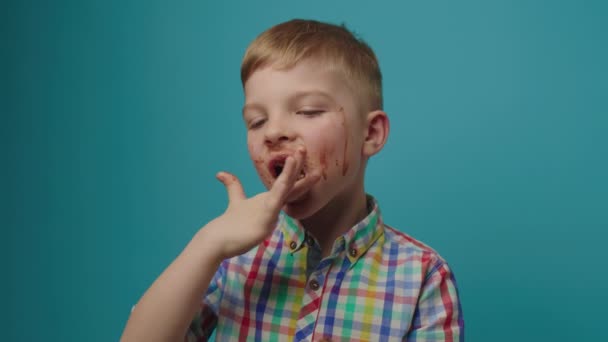 Niño feliz con la cara sucia comiendo barra de chocolate sosteniendo en las manos sonriendo a la cámara de pie sobre fondo azul. Chico alegre disfrutando de dulces de chocolate. — Vídeos de Stock