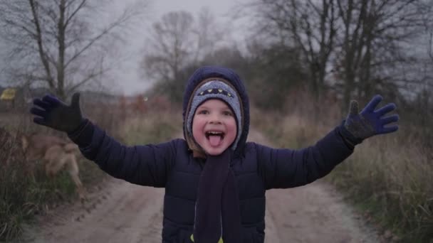 Feliz niño preescolar haciendo caras graciosas mirando a la cámara y huyendo en el sombrío bosque de otoño. Clima frío lluvioso otoño. — Vídeos de Stock