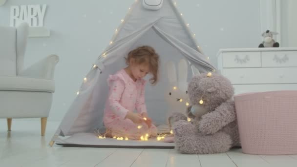 Menina pré-escolar em pijama rosa brincando com ursinho de pelúcia e iluminação guirlanda sentado em tenda infantil na sala de crianças. Andar crianças jogos. — Vídeo de Stock