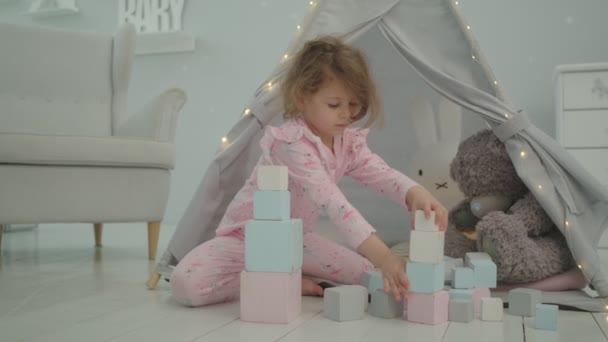 ピンクのパジャマ姿のかわいい女の子が、熊やウサギとテントに座っている木製のブロックで遊んでいます。子供は子供部屋で一人で遊ぶ. — ストック動画