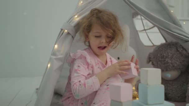 분홍색 파자마를 입은 귀여운 소녀가곰 이랑 토끼 랑 텐트에 앉아 나무 블록을 가지고 노는 것을 보 세요. 아이들은 아이들 방에서 혼자 노는데. — 비디오
