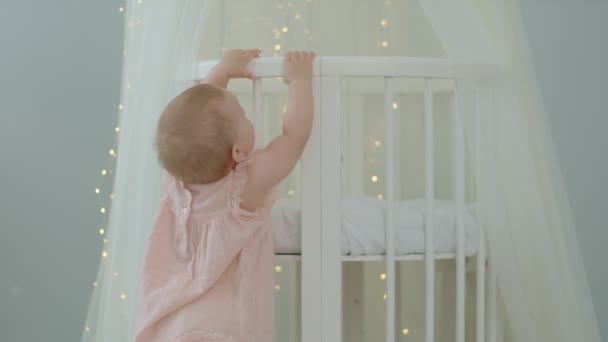 Liten flicka i rosa klänning lära sig att gå håller barnen säng. Första stegen i ljust rum. — Stockvideo