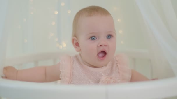 Close up of wondering 10 months old baby girl with blue eyes sitting in bed with canopy with lights. Tender and bright babies room with infant in bed. — Wideo stockowe