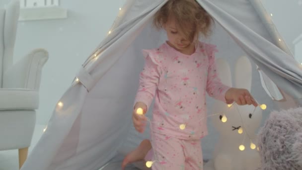 Preschool girl in pink pajama playing with lighting garland sitting in child tent in kids room. Floor kids games. — ストック動画