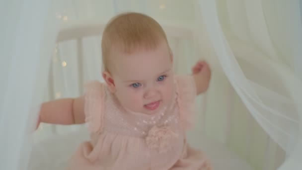 Närbild 10 månader gammal flicka med blå ögon sitter i sängen med krontaket med ljus. Tender och ljusa spädbarn rum med spädbarn i sängen. — Stockvideo