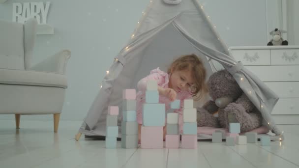 Niña en pijama rosa jugando con bloques de madera sentados en tienda de campaña para niños con oso y conejo. Niño juega solo en la habitación de los niños. — Vídeos de Stock