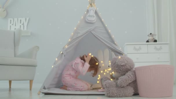 Νηπιοκόμος σε ροζ πιτζάμα παίζει με αρκουδάκι και φωτισμό γιρλάντα κάθεται σε παιδική σκηνή στο παιδικό δωμάτιο. Παιδικά παιχνίδια στο φωτεινό δωμάτιο. — Αρχείο Βίντεο